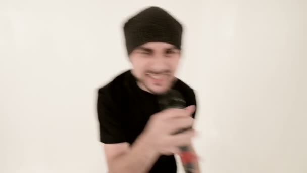 Frontmann Sänger Rockpop mit stylischem Bart in schwarzer Kleidung und Hut mit Mikrofon in den Händen singt ausdrucksstark aggressiv im Studio vor dem Hintergrund weißer Wände — Stockvideo