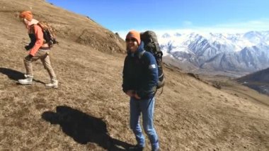 Şapka lı ve güneş gözlüklü sırt çantaları olan iki gülen turist fotoğrafçısı, ellerinde kameralarla sarı çimenlerin üzerindeki tepeye karlı dağların arka planına doğru çıkıyor. yavaş çekim