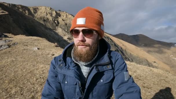 Retrato de um viajante barbudo em óculos de sol e um boné se senta em uma rocha contra o pano de fundo das montanhas. Rindo — Vídeo de Stock