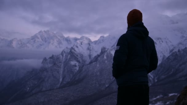 Retrato de um jovem homem barbudo viajante ao anoitecer nas montanhas. Fique pensando seriamente e olhando ao redor. Cara calorosamente vestido no fundo de montanhas nevadas — Vídeo de Stock