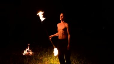 Düşük anahtar. Uzun saçlı ve çıplak gövdeli genç bir erkek, yanan bir meşaleyi alevlerin yanında siyah bir gece videosu yla açık havada döndürür. Modern fakir yanan bir personel ile hileler yapar