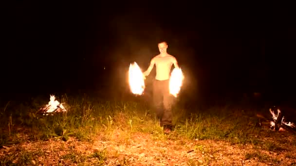 低键。一个留着长发和赤裸躯干的年轻男性在火场附近的黑色夜间视频中旋转燃烧的火炬。现代法基尔做技巧与燃烧的工作人员 — 图库视频影像