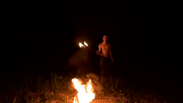 Très discret. Un jeune homme aux cheveux longs et au torse nu tourne une torche brûlante à l'extérieur sur une vidéo de nuit noire au ralenti près des feux. Fakir moderne fait des tours avec un bâton brûlant — Video