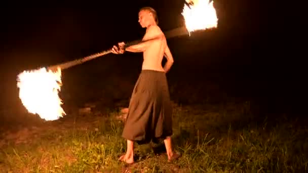 Низко. Молодой мужчина с длинными волосами и обнаженным туловищем вращает горящий факел на улице на чёрном ночном видео, замедленном движении возле костра. Современный факир делает трюки с жгучим посохом — стоковое видео