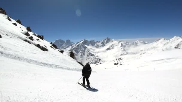 Uno sciatore maschio grandangolare invecchiato in attrezzatura nera e casco bianco con racchette da sci cavalca su un pendio innevato in una giornata di sole. Il concetto di sport sciistici invernali — Video Stock