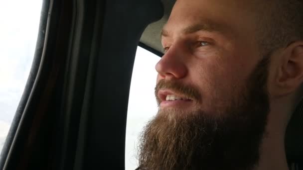 Κοντινό-up πορτραίτο ενός άντρα που μιλάει με γένια, που κάθεται στο αυτοκίνητο στο πίσω κάθισμα και διαλογίζεται στις πλευρές με φόντο τα βουνά — Αρχείο Βίντεο
