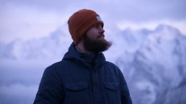 Dağlarda alacakaranlıkta genç sakallı bir erkek gezginin portresi. Ayakta düşünmek ve mantığa gülümseyerek. Karlı dağların arka planda sıcak giyimli adam