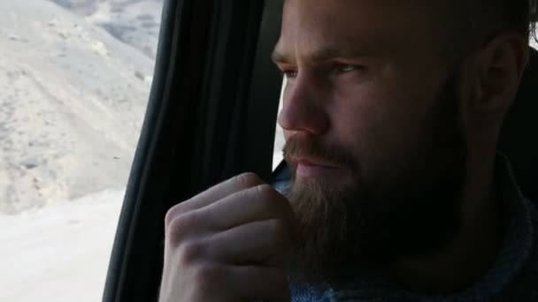 Retrato de close-up de um homem hipster barbudo falando sentado no carro no banco de trás e meditando nas laterais contra o fundo das montanhas — Vídeo de Stock