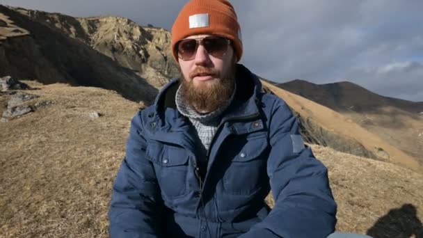 Retrato de un viajero barbudo con gafas de sol y una gorra sobre una roca sobre el telón de fondo de las montañas. Dice que se frota las manos — Vídeo de stock