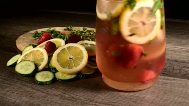 Primo piano in una caraffa di vetro sono fette di limoni e cetrioli con menta e fragole congelate che vengono mescolate in acqua frizzante. Frutta rotante in un bicchierino di limonata — Video Stock