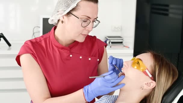 Menina loira bonita em óculos protetores amarelos no stamotologist examinou sua boca aberta. Dentista feminina examina a cavidade oral de um paciente jovem com a ajuda de um instrumento de dentista — Vídeo de Stock