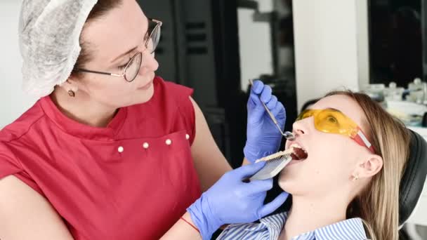 Bella ragazza bionda con occhiali gialli protettivi sulla stamotologa ha esaminato la bocca aperta. La dentista femminile esamina la cavità orale di un giovane paziente con l'aiuto di uno strumento dentista — Video Stock