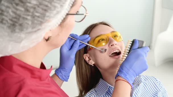 Linda chica rubia con gafas protectoras amarillas en el estamotólogo examinó su boca abierta. Dentista mujer examina la cavidad oral de una paciente joven con la ayuda de un instrumento de dentista — Vídeo de stock