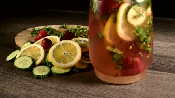 Nahaufnahme in einer Glaskaraffe sind Zitronen- und Gurkenscheiben mit Minze und gefrorenen Erdbeeren, die in sprudelndem Wasser gerührt werden. rotierende Früchte im Limonadenglas — Stockvideo