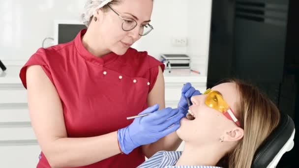 Ganska blond flicka i skyddande gula glasögon på stamotologen undersökte hennes öppna mun. Kvinnlig tandläkare undersöker munhålan hos en ung patient med hjälp av en tandläkare instrument — Stockvideo