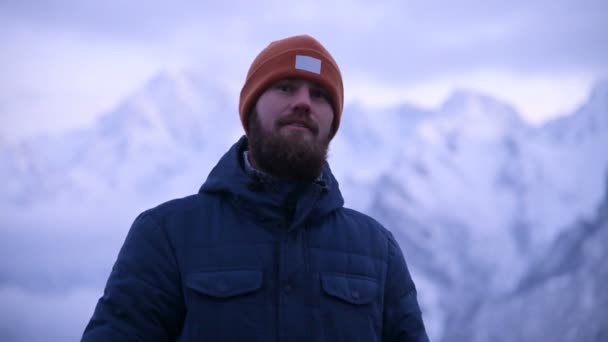 Retrato de un joven viajero barbudo en el crepúsculo en las montañas. Pensar de pie y sonreír a la razón. Tipo bien vestido en el fondo de las montañas nevadas — Vídeo de stock