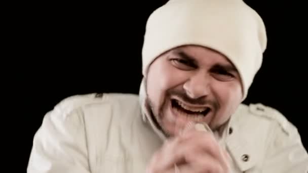 クローズアップフロントマンのボーカリストロックポップは、白い服を着たスタイリッシュなひげと、黒い壁を背景にスタジオで積極的に歌うマイクを手にした帽子 — ストック動画
