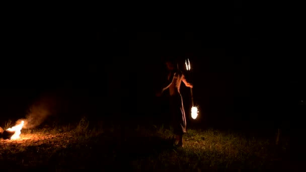 Low key. A young male with long hair and a naked torso rotates a burning torch outdoors on a black night video slow motion near the fires. Modern fakir does tricks with a burning staff — Stock Video