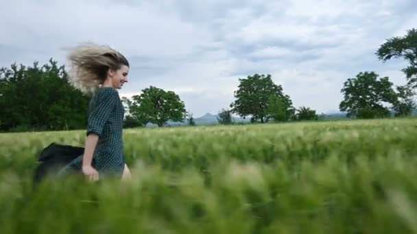 Genç mutlu sarışın kız yağmur gökyüzünün arka plan karşı akşam yeşil bir buğday tarlasında çalışır. Yan görünüm — Stok video