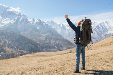 Şapkalı ve sırt çantasıolan sakallı bir adam güneşli bir günde karlı dağ zirvelerine karşı dışarıda duruyor. Arkadan bak. Eller kadar zafer jesti
