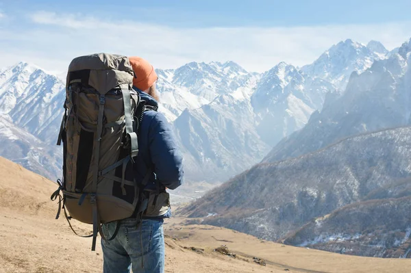 Un uomo barbuto con un cappello e uno zaino è in piedi all'aperto contro le cime delle montagne innevate in una giornata di sole. Vista posteriore — Foto Stock