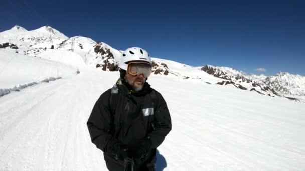 Um esqui macho de grande ângulo selfie envelhecido em equipamento preto e passeios de capacete branco em uma encosta nevada em um dia ensolarado. O conceito de esportes de esqui de inverno — Vídeo de Stock