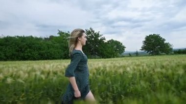 Genç mutlu sarışın kız yağmur gökyüzünün arka plan karşı akşam yeşil bir buğday tarlasında çalışır. Yan görünüm