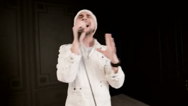 Frontman zpěvák se stylovým plnovousem v bílých šatech a klobouk s mikrofonem v dlaních velmi agresivně zpívá ve studiu na pozadí černých zdí — Stock video