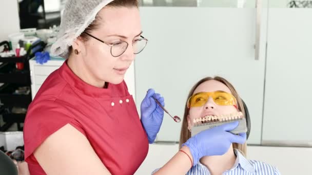 Mooi blond meisje in beschermende gele bril op de stamotoloog onderzocht haar open mond. Vrouwelijke tandarts onderzoekt de mondholte van een jonge patiënt met behulp van een tandarts-instrument — Stockvideo