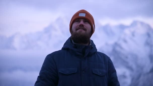 Портрет молодого бородатого путешественника в сумерках в горах. Стоя, размышляя и улыбаясь разуму. Тепло одетый парень на фоне снежных гор — стоковое видео