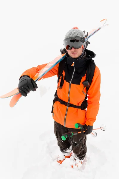 Retrato de un atleta esquiador profesional con un sombrero de punto y un traje negro anaranjado con una máscara de esquí negra con esquís en el hombro durante una tormenta de nieve sobre un fondo claro en la nieve. Ángulo ancho —  Fotos de Stock