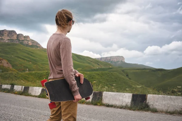 Uzun saçlı genç şık adam bir at kuyruğu ve güneş gözlüğü içinde destansı kayaların arka planda dağlarda bir ülke asfalt yolda elinde bir longboard ile duruyor