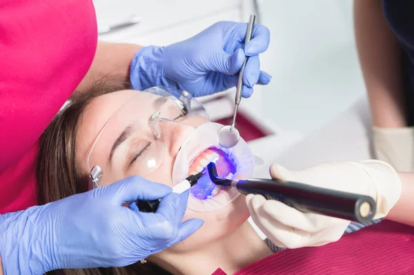 Κοντινό πλαίσιο ενός οδοντιάτρου και νοσοκόμου που φτιάχνει μια αποξηραμένη νέα γέμιση με υπεριώδης νεαρή γυναίκα ασθενή στο οδοντιατρείο — Φωτογραφία Αρχείου