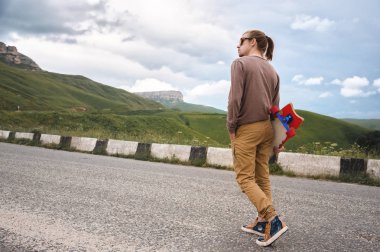 Uzun saçlı ve güneş gözlüğü ile genç şık hipster adam destansı kayaların arka planda dağlarda bir ülke asfalt yolda elinde bir longboard ile yürüyüş