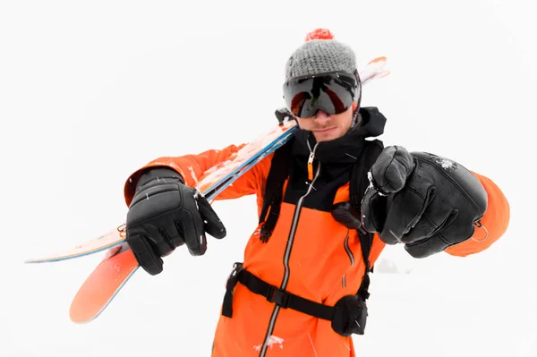 Atleta profissional esquiador em um chapéu de malha e terno preto laranja com uma máscara de esqui preto com esquis no ombro aponta para a câmera durante uma tempestade de neve em um fundo leve na neve . — Fotografia de Stock