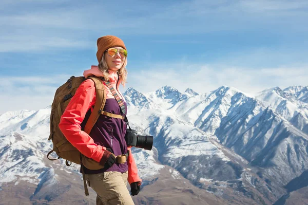 Portré fotós csípő turista lány egy kamera napszemüveg és kalap ellen, a háttérben a havas Kaukázus hegység. Fényképes utazási koncepció — Stock Fotó