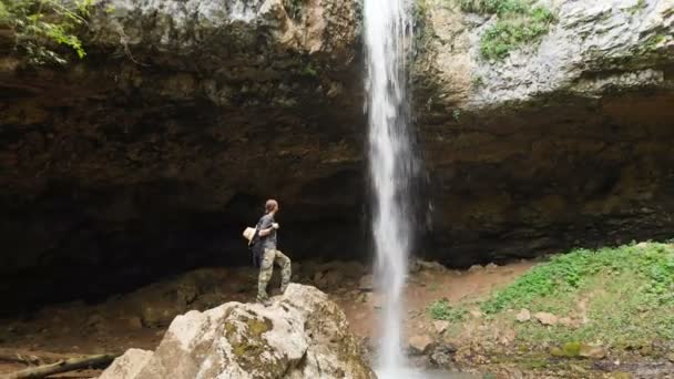 Luchtfoto van een mannelijke reiziger met een rugzak staande op de rotsen voor een majestueuze waterval en het bewonderen van de natuur in het bos tussen de rotsen. Reizen video 4k — Stockvideo