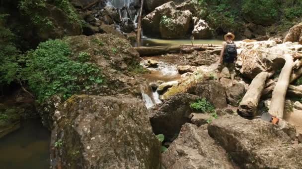 Veduta aerea di un viaggiatore di sesso maschile con un cappello di paglia e uno zaino si arrampica su una roccia sullo sfondo di una maestosa cascata nel bosco tra le rocce. Video di viaggio 4k — Video Stock