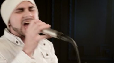 Yakın çekim Frontman vokalist rock pop beyaz giysiler içinde şık bir sakal ve elinde mikrofon ile bir şapka ile ifade agresif siyah duvarların arka plan karşı stüdyoda şarkı