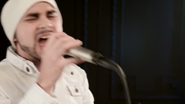 Close-up Frontman vocalista rock pop com uma barba elegante em roupas brancas e um chapéu com um microfone em suas mãos cantando expressivamente agressivamente no estúdio contra o fundo das paredes pretas — Vídeo de Stock