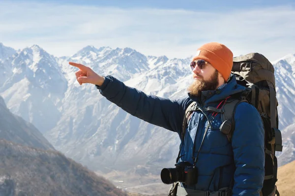 Homme barbu en lunettes de soleil et un chapeau avec un sac à dos pointe la main vers les montagnes. Concept de voyage sur fond de paysage de montagne — Photo