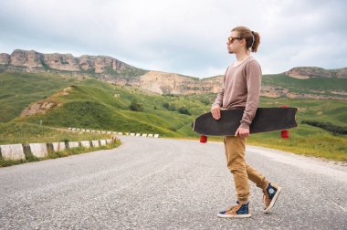 Uzun saçlı ve güneş gözlüğü ile genç şık hipster adam destansı kayaların arka planda dağlarda bir ülke asfalt yolda elinde bir longboard ile yürüyüş