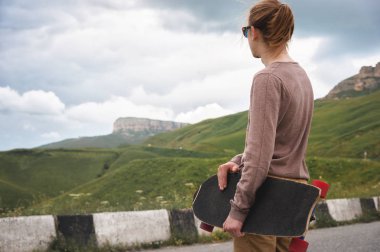 Şık bir genç adam gün batımından sonra akşam elinde bir paten veya longboard ile dolambaçlı bir dağ yolu boyunca ayakta. Gençlik spor ve seyahat hobileri kavramı