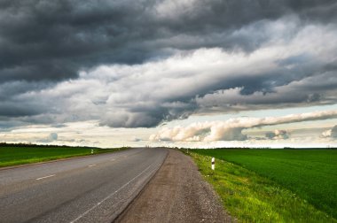 Yanlarda düşük dokulu dramatik bulutlar ve yeşil alanlar ile banliyö asfalt yol Akşam manzara