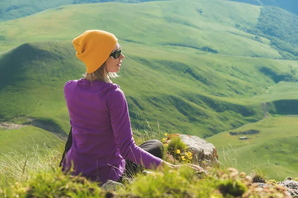 Vista trasera de una atractiva turista sentada en una colina cubierta de hierba con gafas de sol. Disfruta de un día soleado de verano en el Cáucaso. Concepto de turismo de vacaciones activo — Foto de Stock