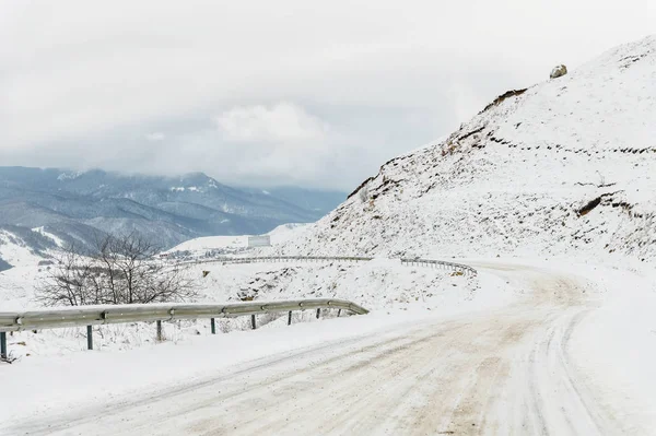 Camino de asfalto de montaña vacío en invierno cubierto de nieve en un día nublado. El concepto de conducir un coche en el hielo de invierno y viajar — Foto de Stock