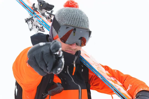 Professional skier athlete in an orange black suit with a black ski mask with skis on his shoulder points to the camera on a light background in the snow