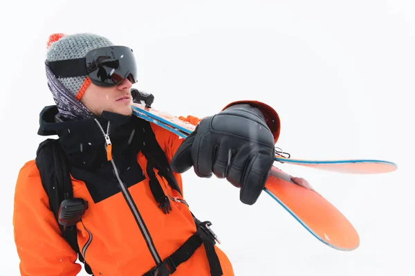 Ritratto di atleta professionista in giacca arancione con maschera nera e con gli sci sulla spalla guarda di lato. sfondo grigio durante la tempesta di neve — Foto Stock