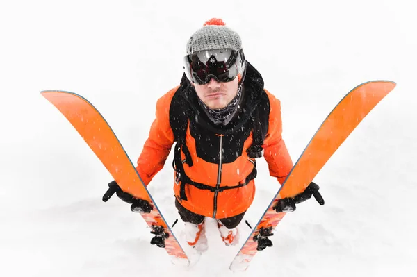 Un atleta esquiador profesional con un traje negro naranja con un pasamontañas negro con esquís en las manos se para con una masa descontenta durante una tormenta de nieve sobre un fondo claro en la nieve —  Fotos de Stock