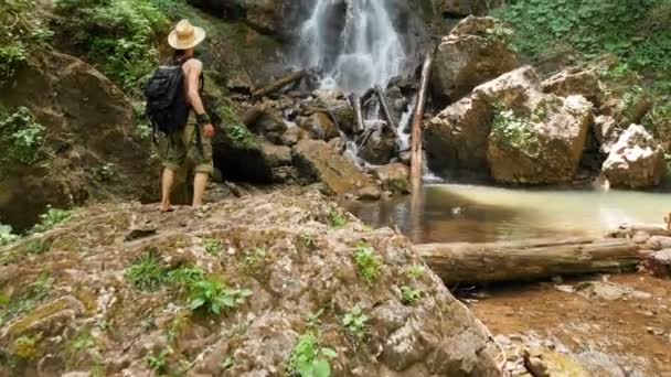 Vista aérea de un viajero masculino en un sombrero de paja y una mochila sube sobre una roca en el fondo de una majestuosa cascada en el bosque entre las rocas. Viajes video 4k — Vídeo de stock